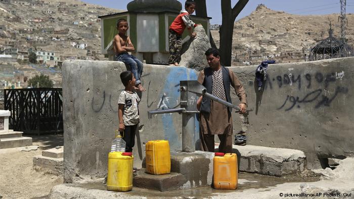 Un hombre y tres niños llenan grandes bidones amarillos de una fuente de agua pública.