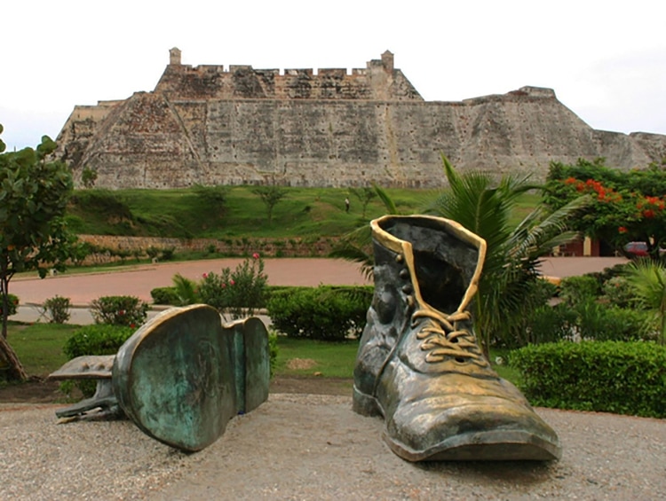 La obra los Zapatos Viejos con el Castillo de San Felipe de fondo. Esta obra es la réplica hecha por Héctor Lombana, hermano de Tito, que demolió la original por esta de bronce y se adjudicó su autoría.