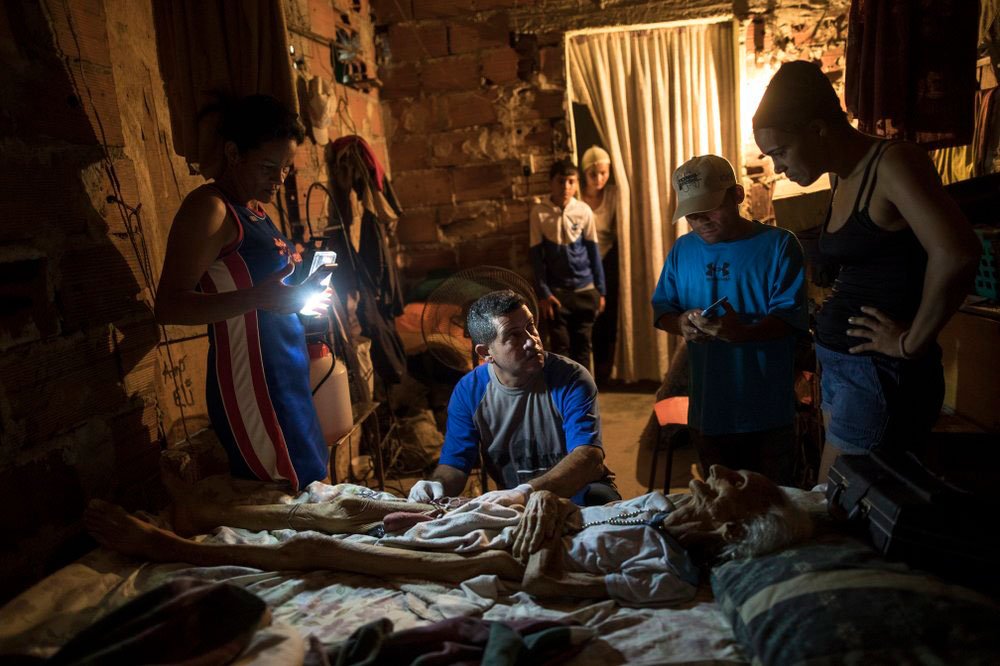 La activista comunitaria Carolina Leal usa un teléfono celular para iluminar el área de trabajo de Roberto Molero mientras prepara el cuerpo de Teresa Jiménez, de 91 años, después de que ella muriera por causas naturales en su casa en Maracaibo, Venezuela, el 17 de noviembre de 2019 (AP)