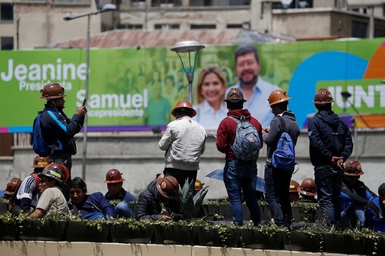 Mineros frente a un cartel de propaganda del binomio Jeanine Anez y Samuel Doria Medina en el centro de La Paz. La campaña está paralizada por la pandemia de coronavirus. (REUTERS/David Mercado)