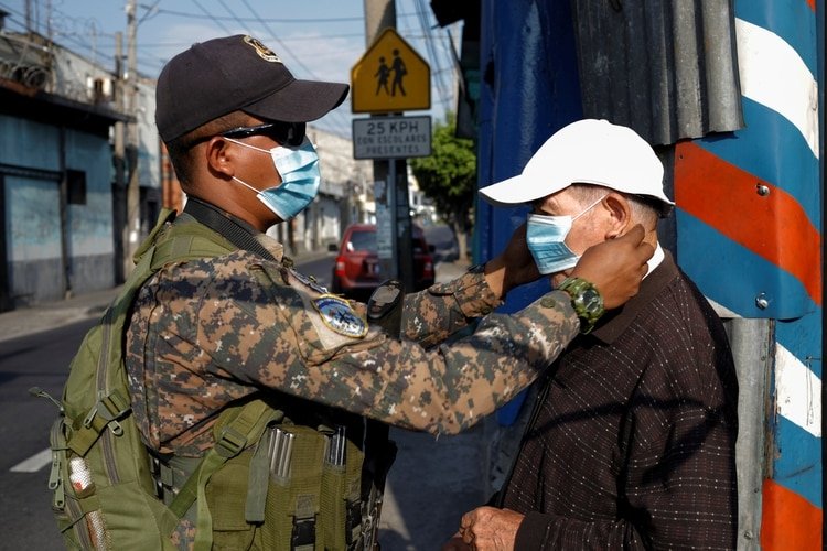 Un soldado ayuda a un hombre a ponerse una máscara facial en un puesto de control en San Salvador (El Salvador) el 20 de abril de 2020. (REUTERS/Jose Cabezas)